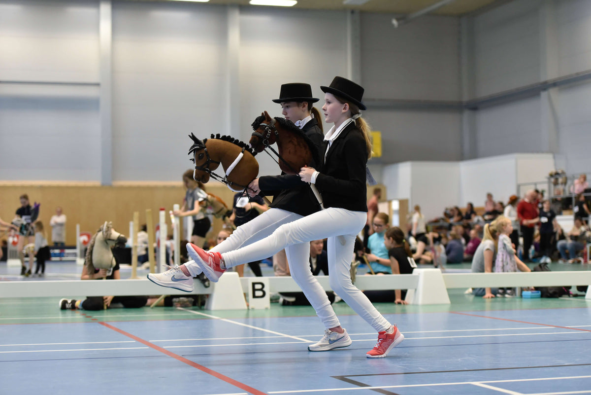 To børn i formelt stævnetøj konkurrerer indendørs med kæpheste i Hobby Horsing