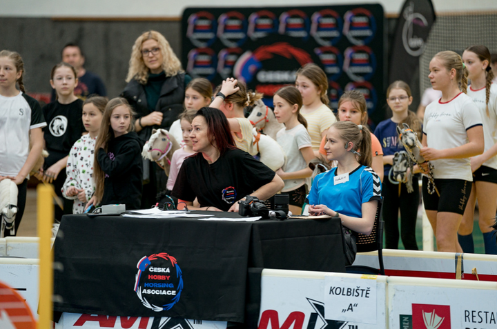 En travl scene fra et indendørs stævne i Hobby Horsing: to arrangører sidder ved et sort bord med logoet for Česká Hobby Horsing Asociace, mens piger i sportstøj står omkring dem med deres kæpheste. Der ses også hvide bander og skiltet »Kolbiště A«.