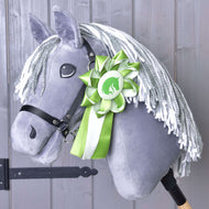 A gray plush Hobby Horse Ribbon Green by Hobby Horse LarDen features a white yarn mane, a black bridle, and a large green and white winning horse ribbon. The wooden stick is visible against a backdrop of wooden paneling.