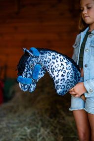 Pige i cowboyjakke i en stald holder en blå og hvid leopardplettet kæphest med den petroleumsblå grime med pels