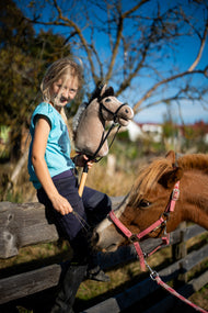 Smilende lyshåret pige på et træhegn med en beige kæphest i sort trense ved siden af en fuksfarvet pony med lyserød grime