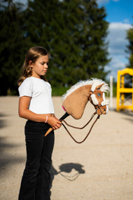 Pige i hvid top på en sandbane holder kæphesten Hobby Horse Star, her med brun grime med cremefarvet pels og brune tøjler af reb