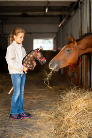 Pige i en stald med en brun, plettet kæphest i vinrød grime med pels ved siden af en brun hest, der spiser hø
