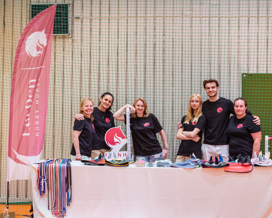 Seks medlemmer af teamet bag LarDen i ens sorte T-shirts med logo poserer bag et bord med farverige tøjler, bånd og tilbehør til Hobby Horsing i en idrætshal. Bag dem ses et lyserødt banner med LarDen Hobby Horses og et net.