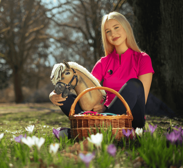 Ung kvinde i en klart lyserød trøje sidder i græsset med en Hobby Horse kæphest og en flettet kurv med farverige æg blandt hvide og lilla blomster