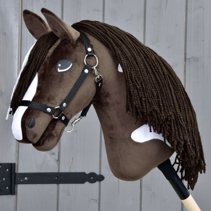 Realistisk brun og hvid Hobby Horse kæphest med sort trense og lang manke af garn på en kæp af træ foran et gråt træhegn