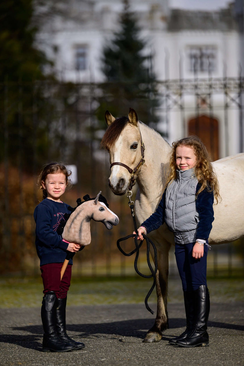 Two young girls stand outdoors beside a white horse; one girl holds a toy hobby horse, reflecting their love for hobby horsing, while the other holds the real horse’s reins. A blurred building and trees can be seen in the background.