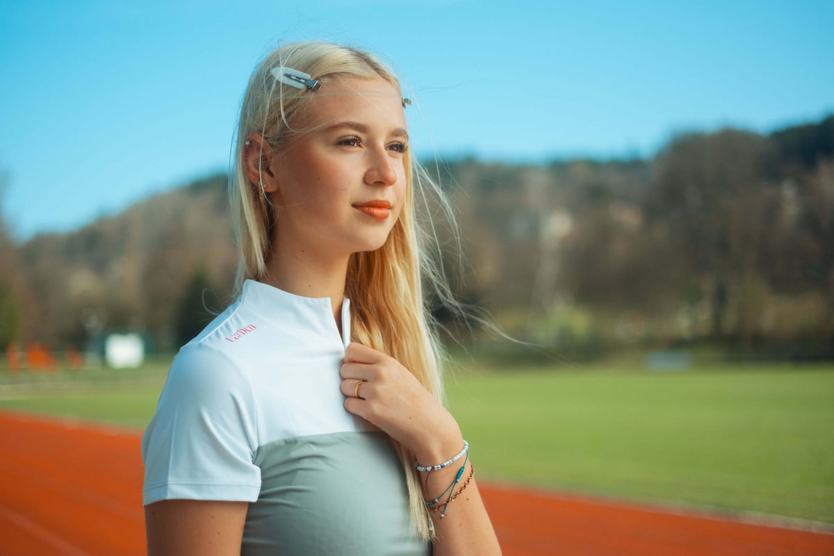 A young blonde woman in a sporty outfit stands on an outdoor running track, thoughtfully holding her hobby horse. The background features green grass, trees, and a blue sky, capturing her passion for hobby horsing.