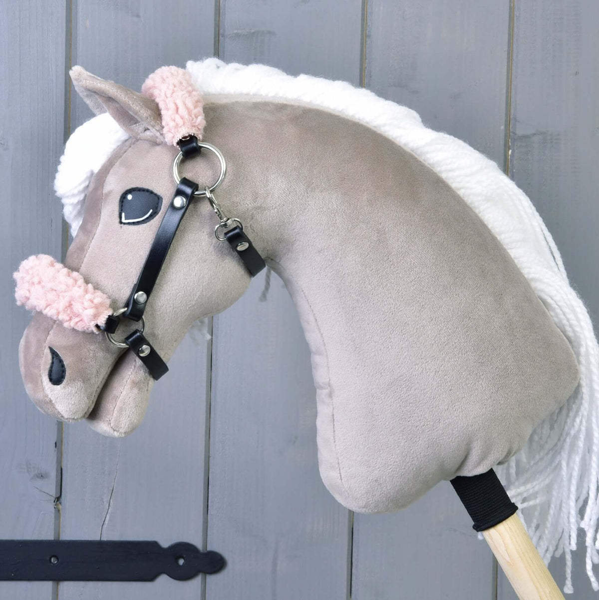 The Hobby Horse LarDen’s Black halter with old rose fur - leatherette features a plush hobby horse with a light brown head, white mane, pink fuzzy bridle, and black leatherette halter on a wooden stick against a gray wooden wall.