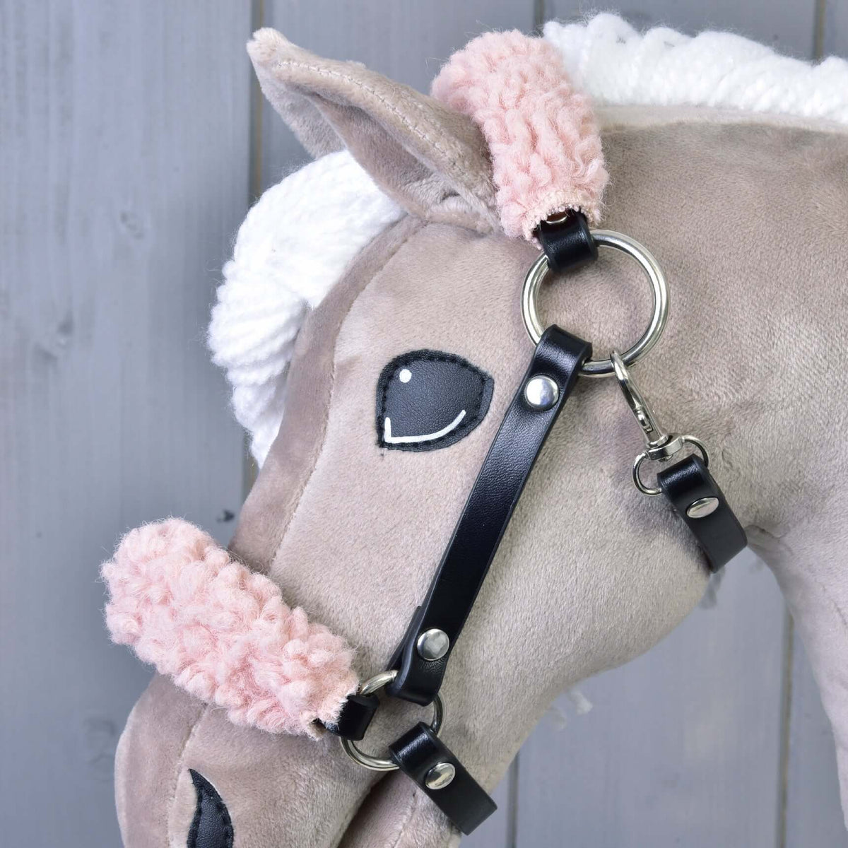A close-up of a plush toy horse’s head with a white yarn mane, black felt eye, and a Hobby Horse LarDen black leatherette halter with old rose fur, set against a light gray wooden background.