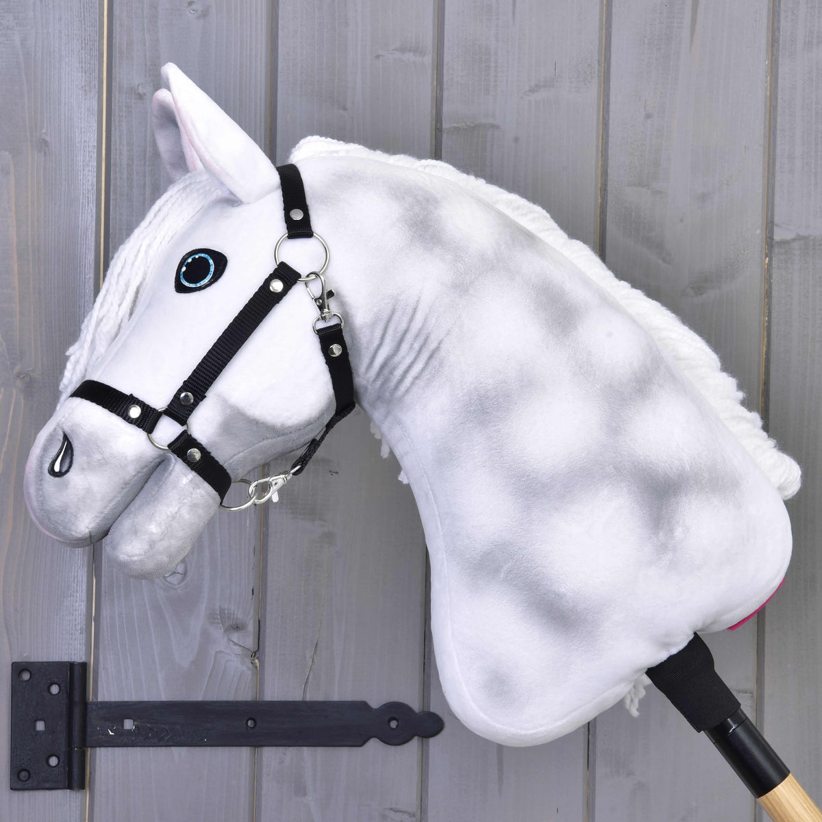 The Hobby Horse LarDen "Elektra" features a plush white design with blue eyes, a black halter, and is mounted on a stick, shown here against a gray wooden wall.