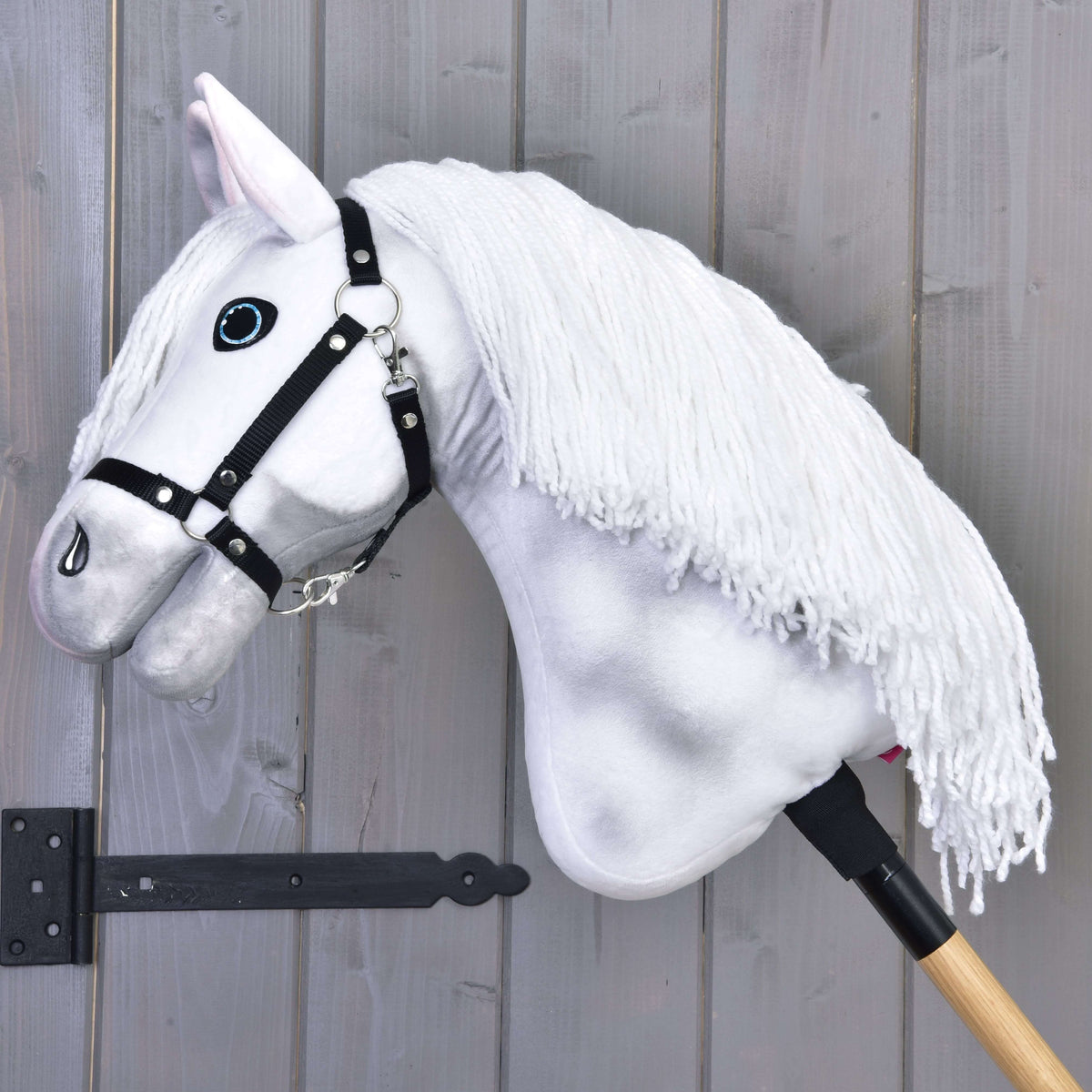 The Hobby Horse LarDen "Hobby Horse Elektra with Black Halter" features a hand-painted white mane, is mounted on a wooden stick, and leans against a gray wooden wall.