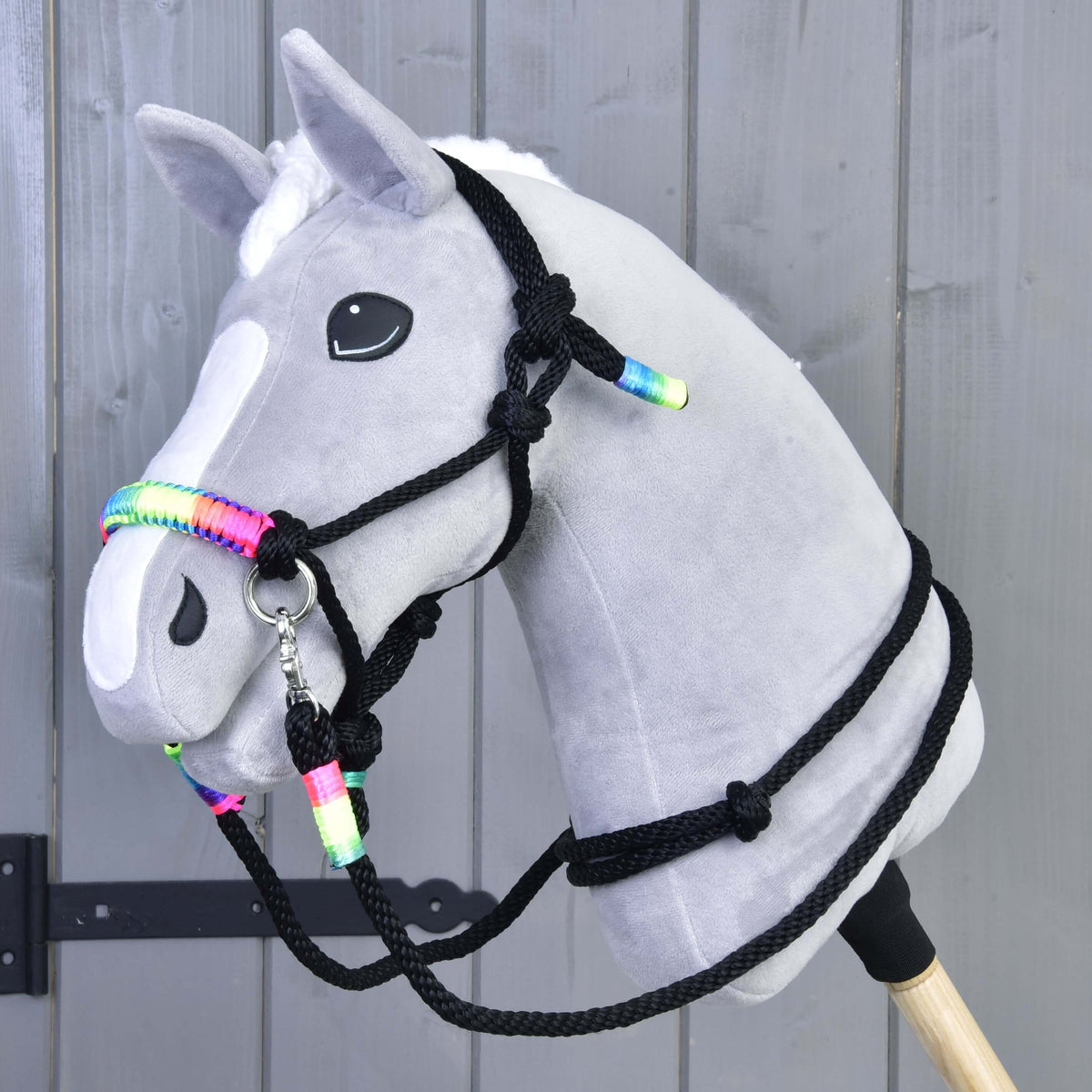 A plush gray hobby horse by Hobby Horse LarDen with a white blaze and mane, wearing the Rope set Rainbow halter, mounted on a wooden stick against a gray wooden panel background.