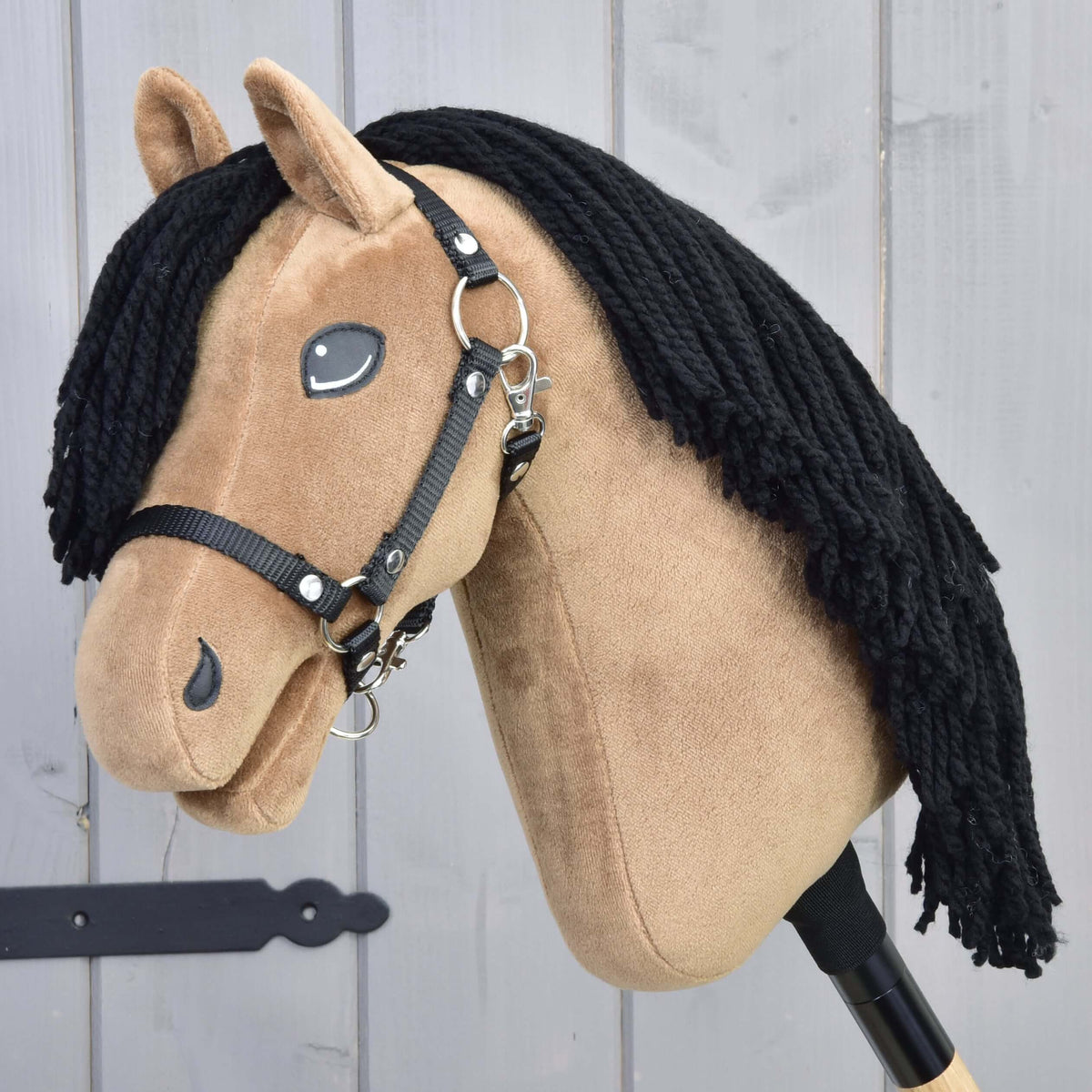 The Hobby Horse LarDen "Hobby Horse Samantha with Black Halter" features a tan fabric head, black yarn mane, and is inspired by an English Thoroughbred mare, shown on a wooden stick in front of gray wood panels.
