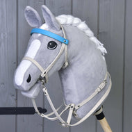 The Hobby Horse LarDen features a plush gray hobby horse with a braided white mane, mounted on a wooden stick, and sporting an Aqua leatherette set—bridle, reins, and martingale—in front of a gray wooden wall.