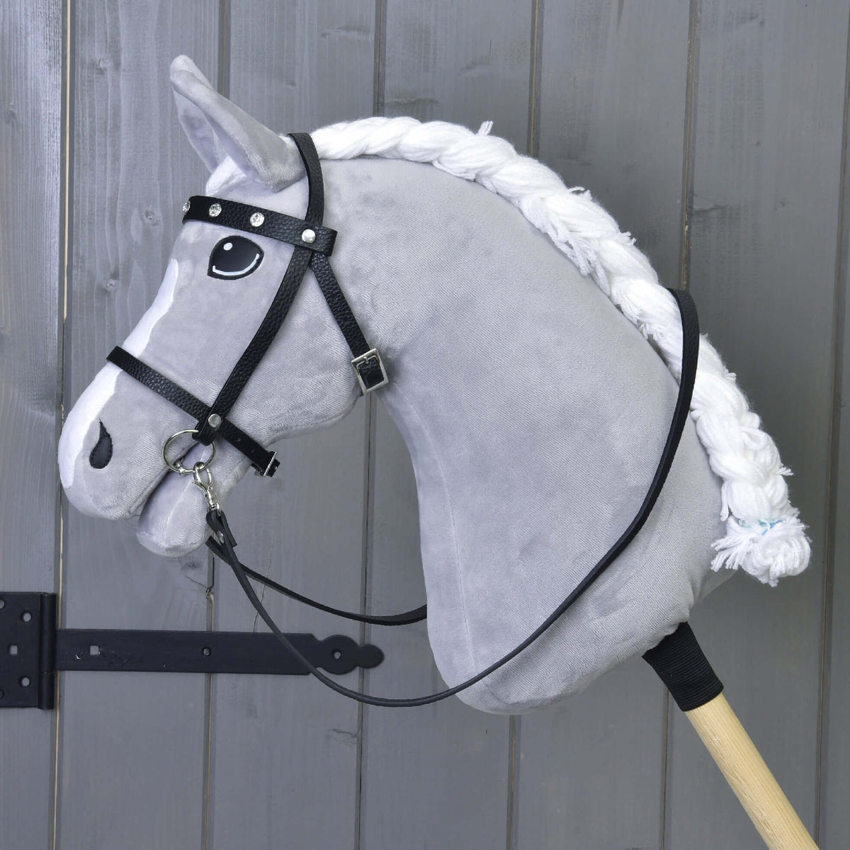 A gray hobby horse with a white mane, wearing Hobby Horse LarDen's complete black bridle with diamonds and detachable reins, is mounted on a wooden stick and set before a wooden wall.