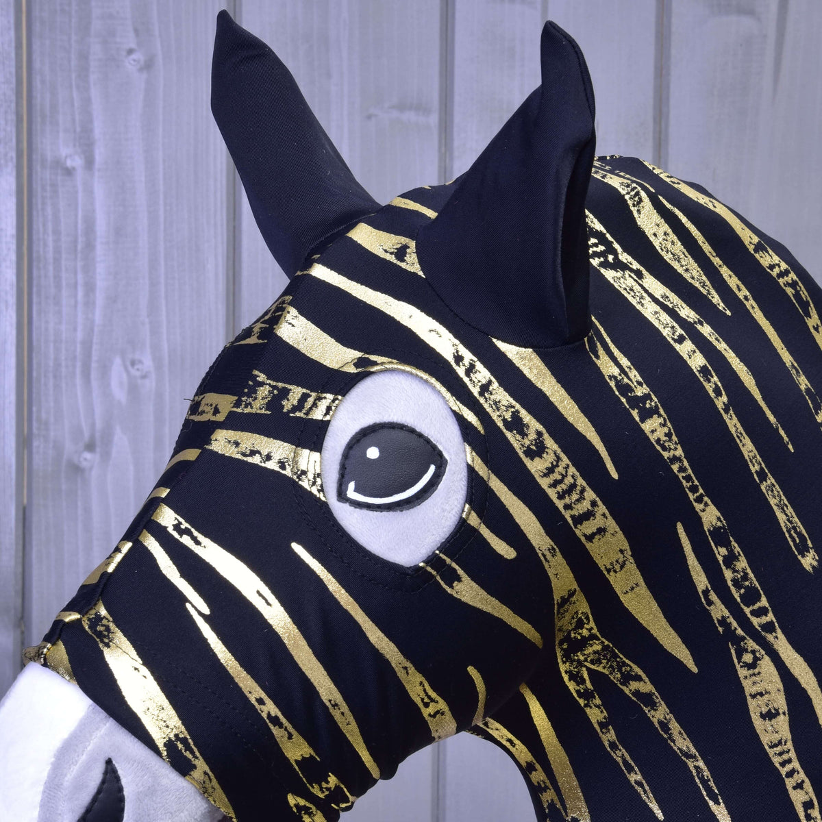 A close-up of the Hobby Horse LarDen protective suit Zebra Gold (Lycra) shows its black and gold striped fabric, large black ears, and cartoonish eye, set against a light wooden background.