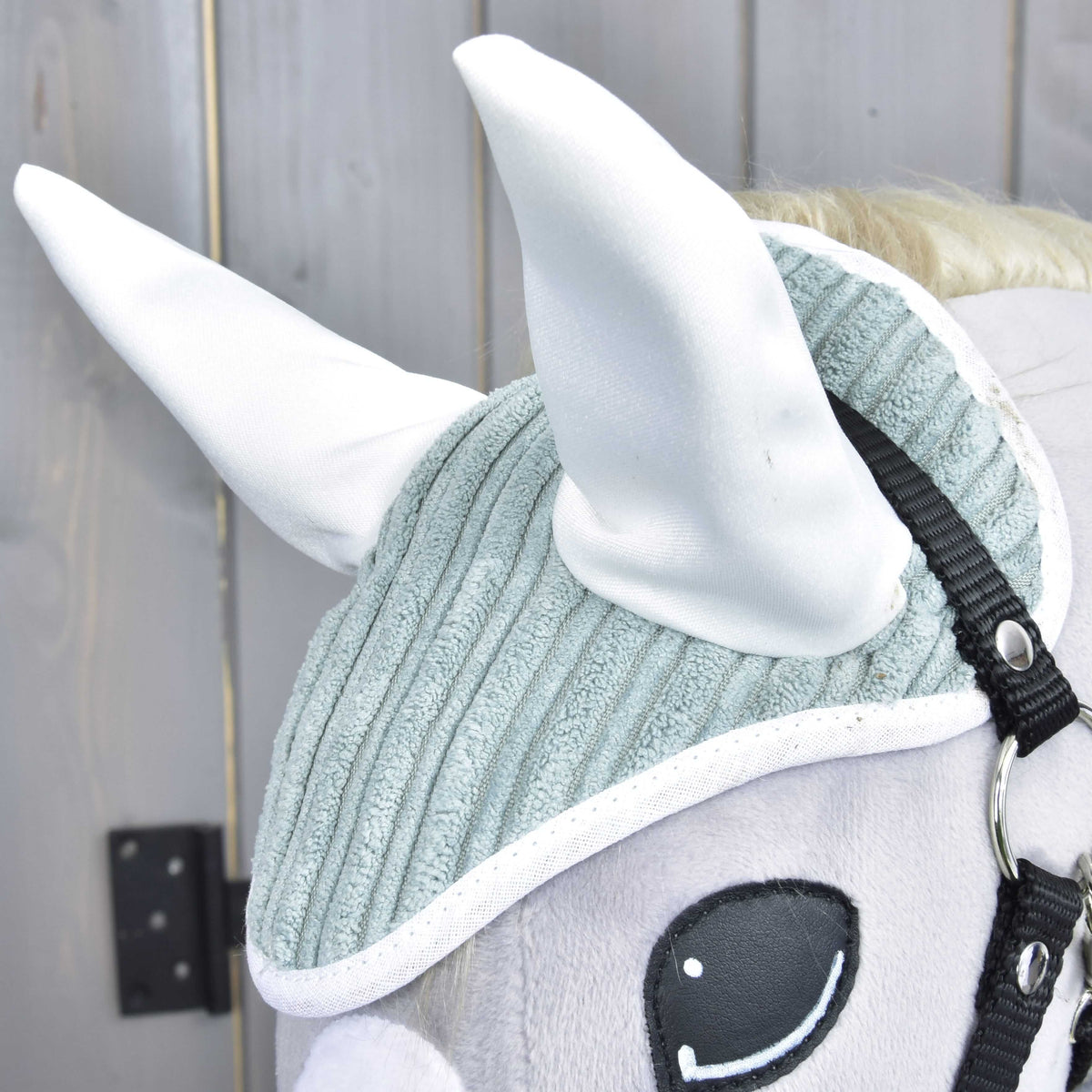 Close-up of Hobby Horse LarDen's Set Manchester Little Green: a plush horse head with a textured light gray ear bonnet, large white ears, black bridle, and embroidered eye, shown against a wooden background.