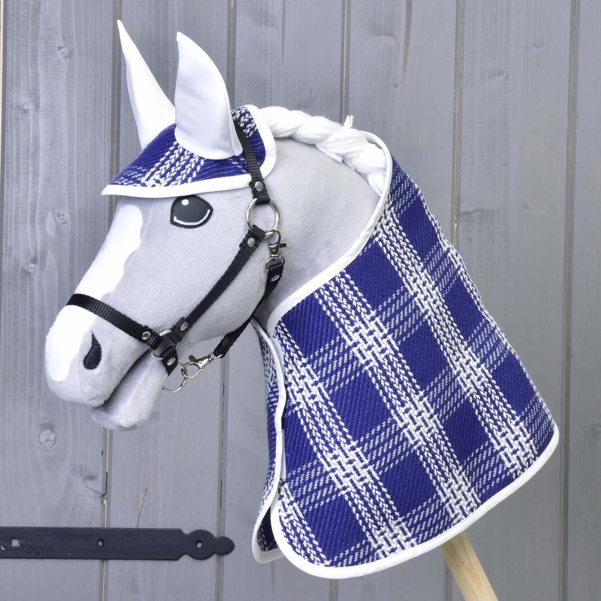A white Hobby Horse LarDen toy with a black bridle wears the Set Blue Cube plaid blanket and cap, standing against a gray wooden background.