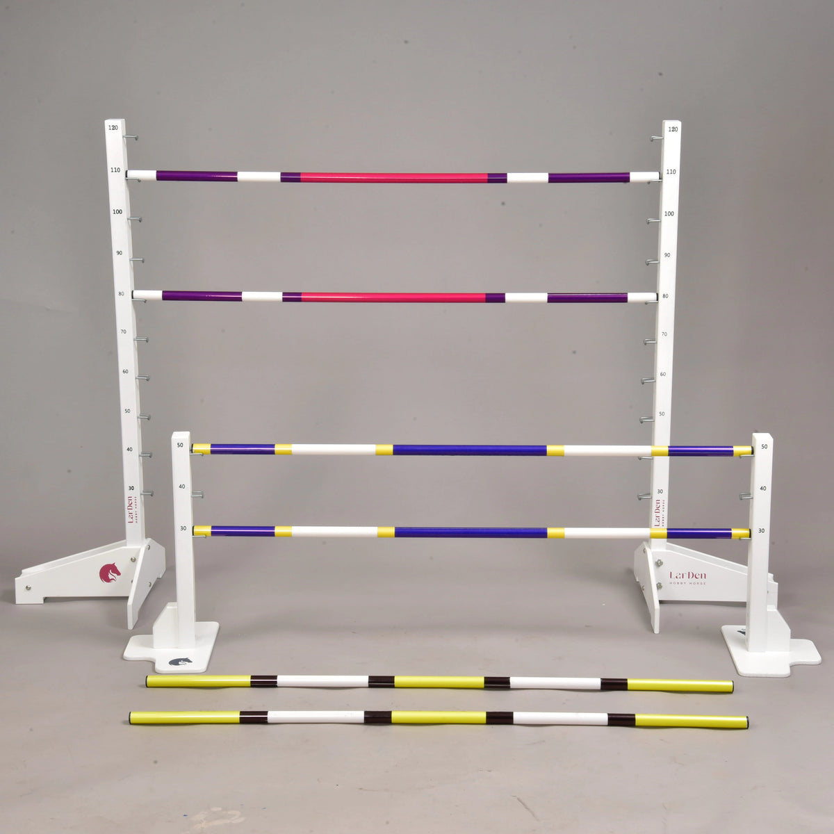 The Hobby Horse LarDen Jump set for Hobby Horsing is arranged in a studio, with adjustable jumps. Some striped purple, yellow, and white poles are set at various heights on stands, while others lie on the ground.