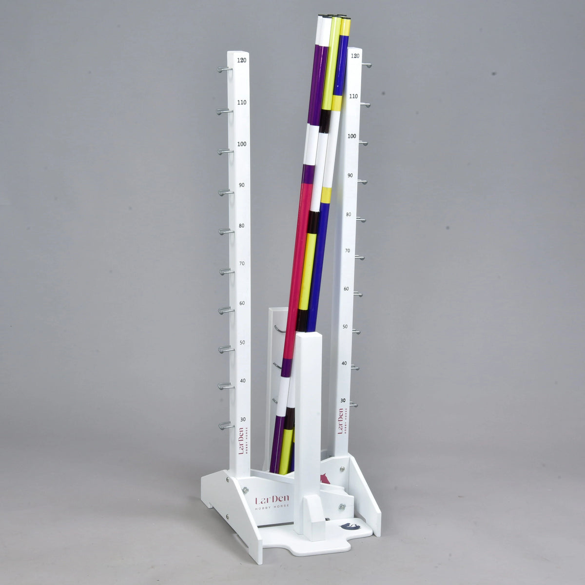 The Hobby Horse LarDen Jump set for Hobby Horsing features a vertical white stand with measurement markings and colorful striped poles in purple, yellow, white, and black—ideal for jumps or adjustable cavaletti. The background is plain gray.