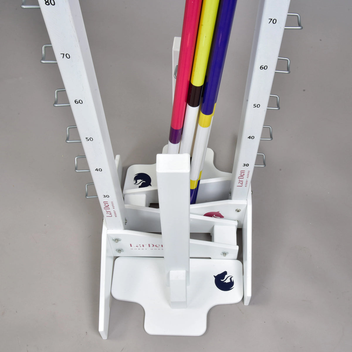 A white vertical rack with numbered markings and hooks holds colorful poles upright for adjustable jumps, ideal for hobby horsing. The base shows a horse logo and the “Hobby Horse LarDen” brand. The floor is light gray.