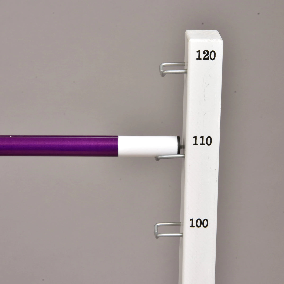 A close-up of the Hobby Horse LarDen Jump set for Hobby Horsing, showing the adjustable bar on a white stand with measurement labels (100, 110, 120), perfect for jumps—set against a plain gray background.