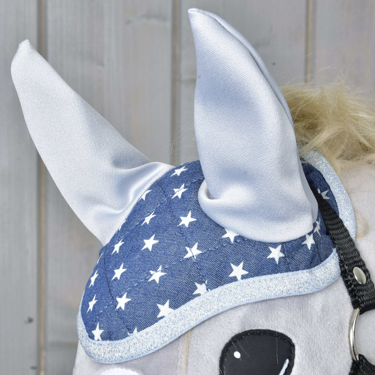 Close-up of a horse wearing the Hobby Horse LarDen Ear bonnet Star, featuring a blue design with white stars and silver trim for stylish fly protection. Part of the horse's eye is visible against a light wood panel background.
