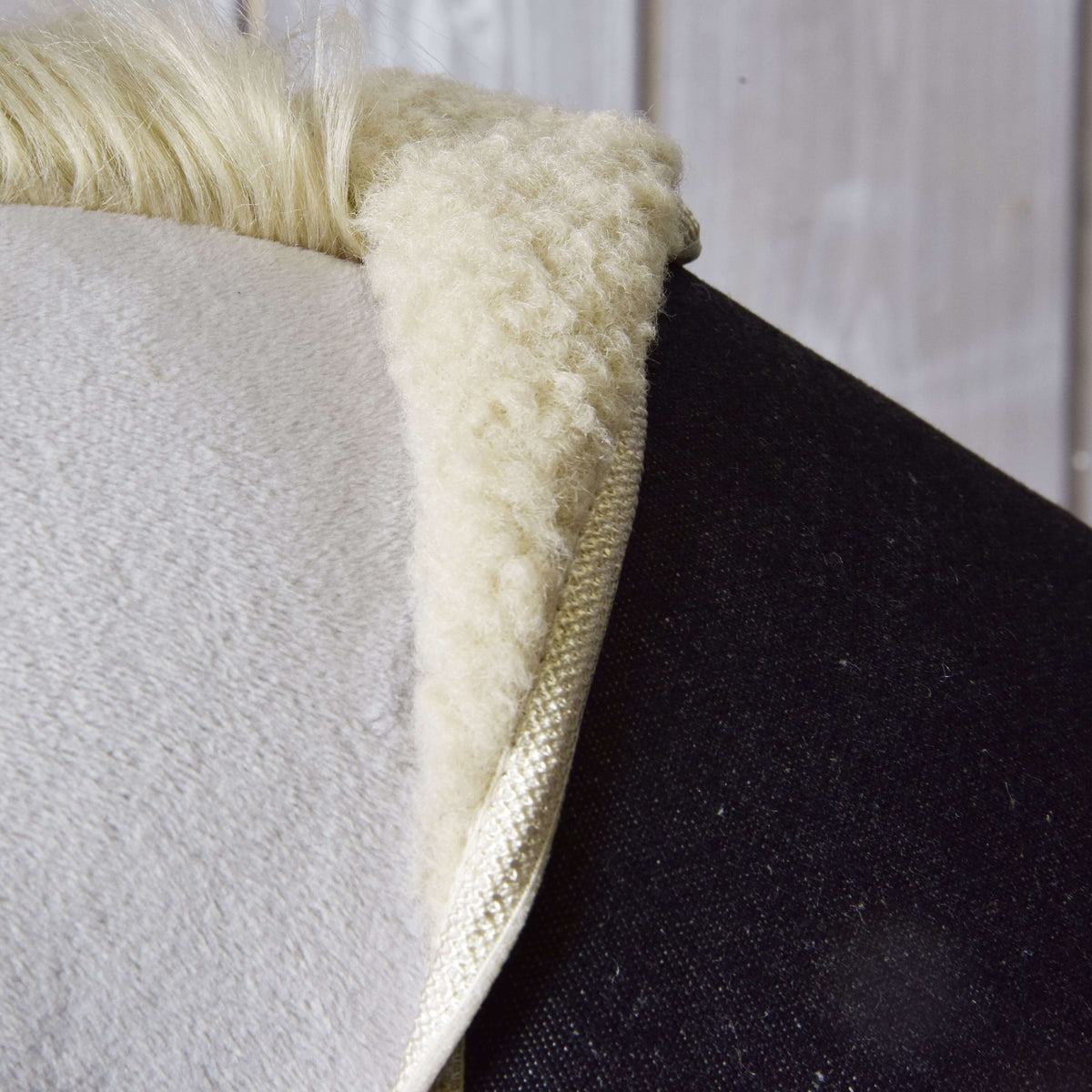 Close-up of the Hobby Horse LarDen "Set Blue Denim" saddle pad, featuring a soft, fluffy white sheepskin texture on a dark horse’s back, with a wooden wall in the background.
