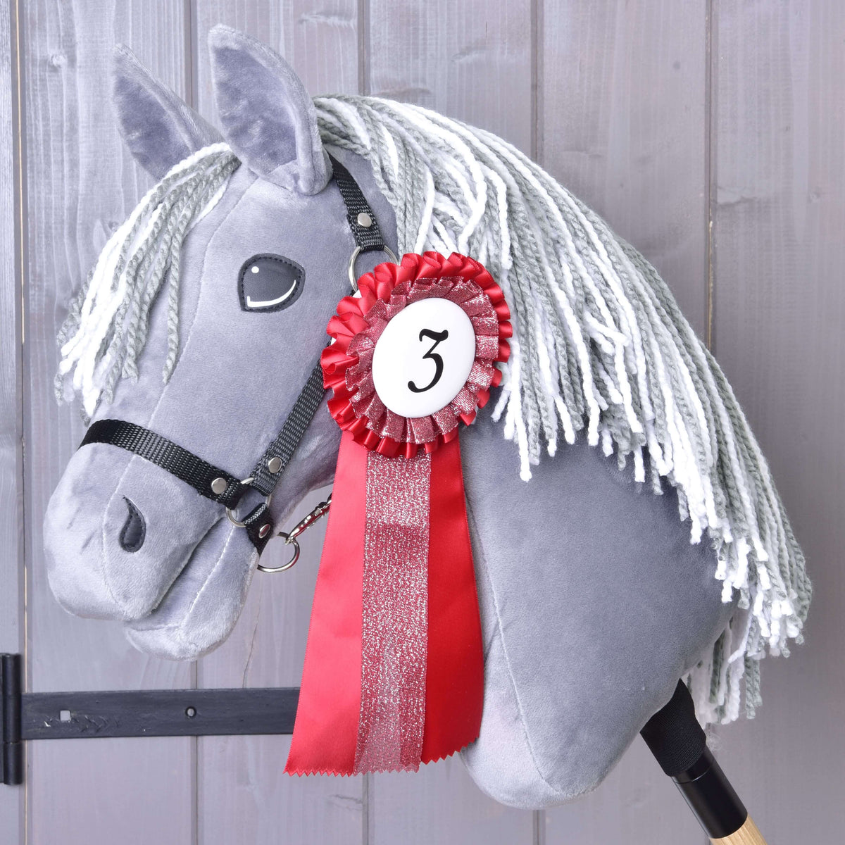 A gray plush hobby horse with a white yarn mane, black bridle, and a red and white Hobby Horse LarDen "Set Hobby Horse Ribbons NEW" pinned with the number 3 stands before gray wooden panels.