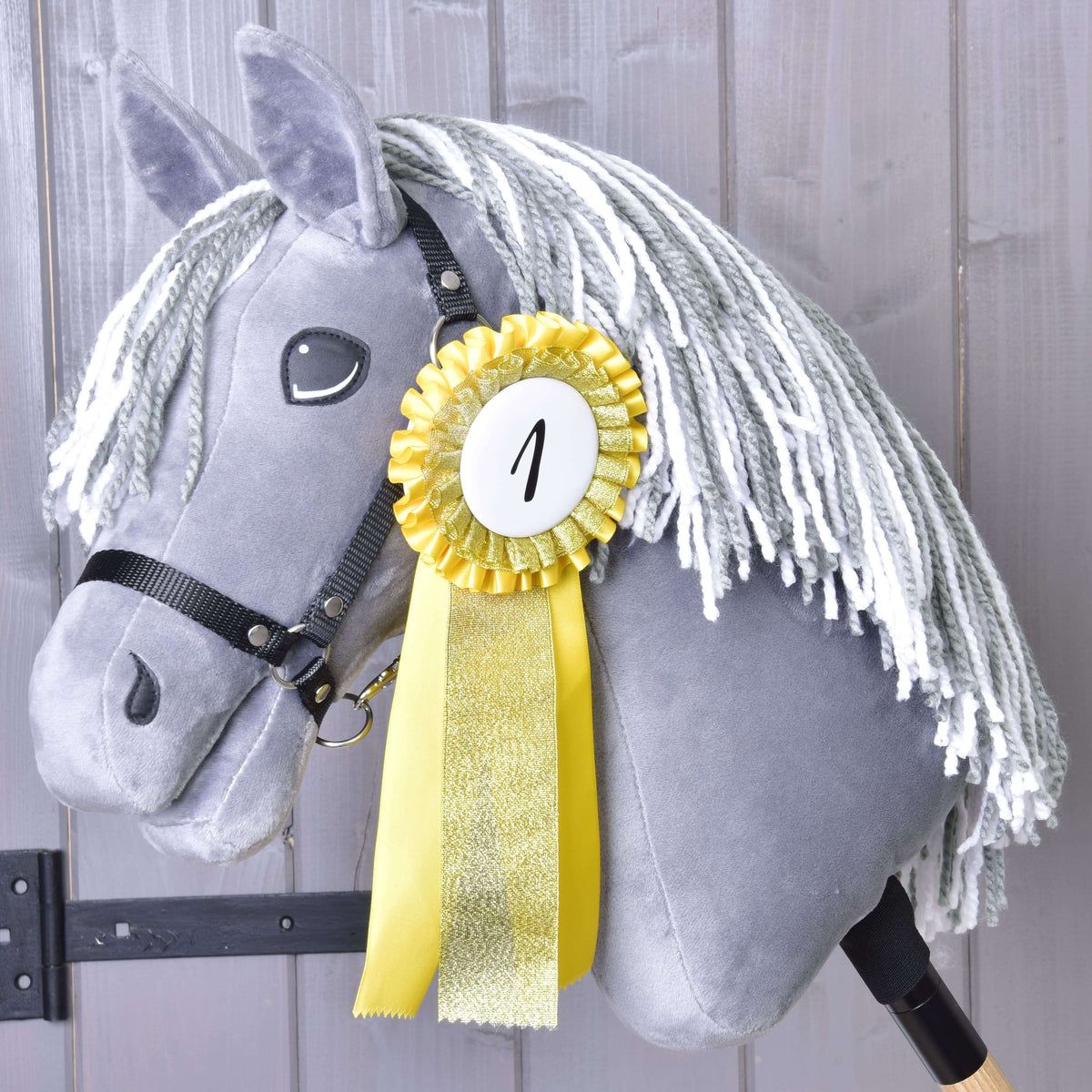 Showcase your achievements with the Set Hobby Horse Ribbons NEW by Hobby Horse LarDen, featuring a yellow first place ribbon marked ‘1’ on a gray hobby horse with white yarn mane, displayed against a wooden wall background.