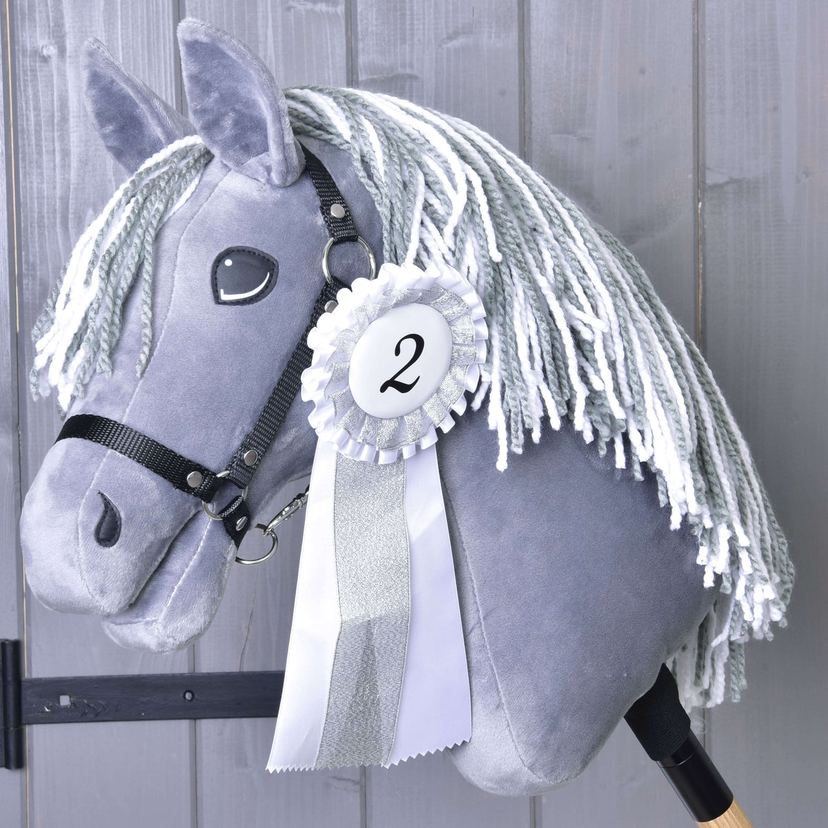 A gray hobby horse with a white yarn mane and black bridle on a wooden stick displays a Set Hobby Horse Ribbons NEW (second place, silver trim, number 2) by Hobby Horse LarDen against gray wooden planks.