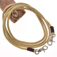 The Hobby Horse LarDen Western Reins Dakota features a tan braided rope with metal snap hooks on both ends, coiled on a wooden branch. The rope ends are wrapped in dark brown material near the hooks.