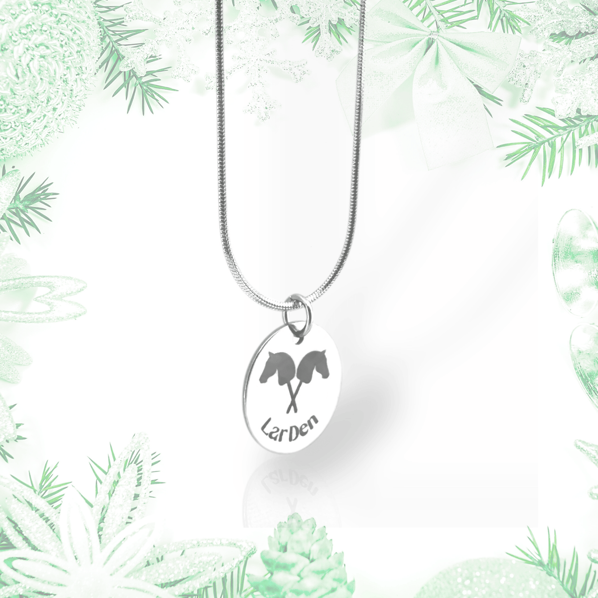 A Necklace with Hobby Horse pendant by Hobby Horse LarDen, crafted from surgical stainless steel, is displayed against a pale green and white festive backdrop with pine branches, bows, and ornaments.