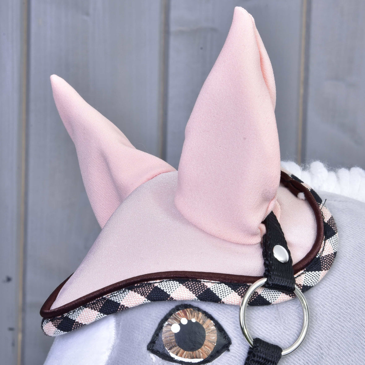 Close-up of the Hobby Horse LarDen Set Abby plush toy wearing a pink ear bonnet with plaid trim and black strap. Large fabric ears and stitched eye are visible against a wooden background.