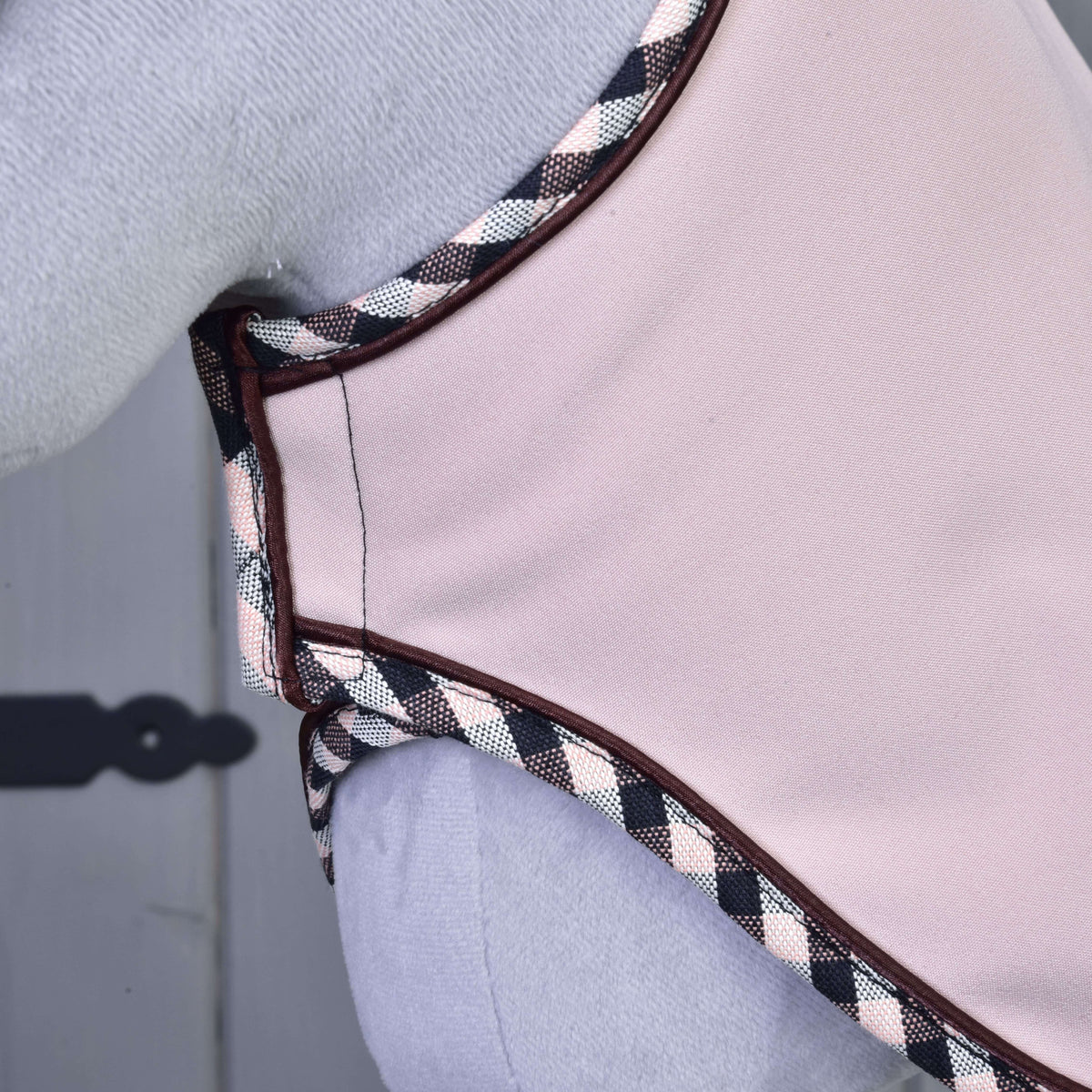 Close-up of Set Abby by Hobby Horse LarDen—a light pink garment with black, white, and pink checkered trim, similar to an ear bonnet—worn against a light background with a decorative hinge visible on the left.