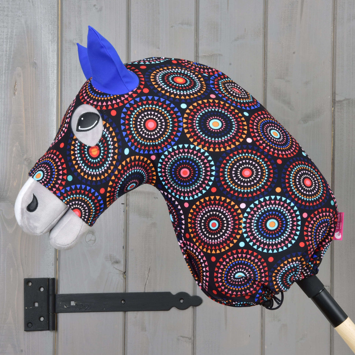 A vibrant hobby horse with a patterned Lycra fabric head, blue felt ears, and white details on a wooden stick—perfectly paired with the Hobby Horse LarDen "Hobby Horse Protective Suit Alice (Lycra)".