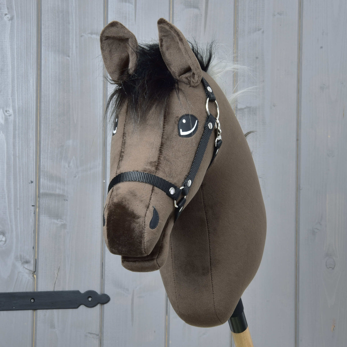 The Hobby Horse LarDen "Hobby Horse Alfredo with Black Halter" features a plush brown English Thoroughbred stallion head with a black mane and bridle, mounted on a wooden stick and set before a gray wooden panel wall.