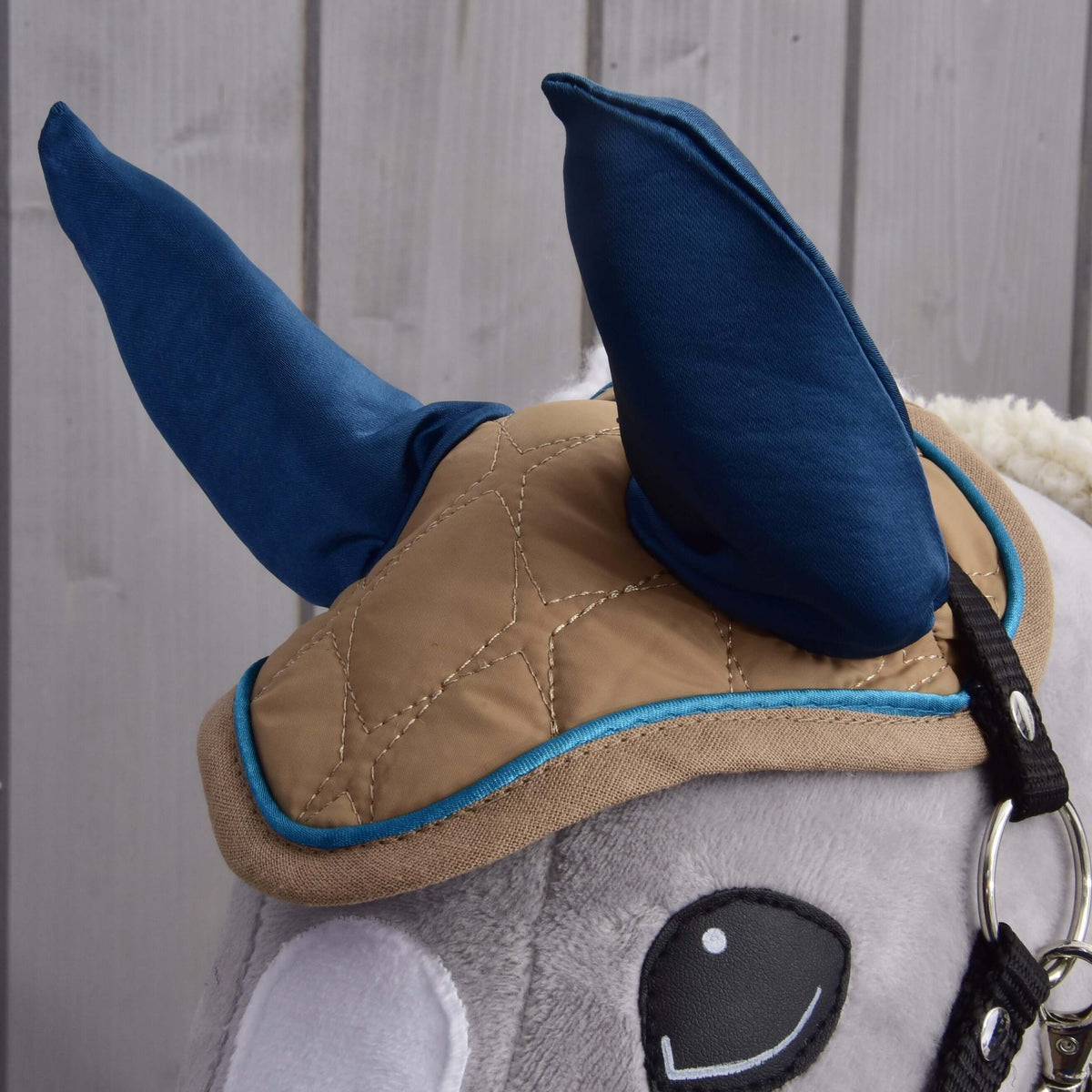 A close-up of the Hobby Horse LarDen Set Almond Star shows its blue ear bonnet, tan quilted blanket with blue trim, and bridle on a plush hobby horse head, set against a light wooden background.