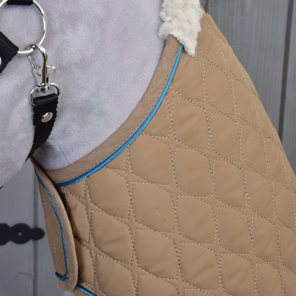 Close-up of the Set Almond Star dog coat by Hobby Horse LarDen, featuring tan quilted fabric with blue trim, fluffy white lining, and secured with a black strap and metal clasp against a wooden background.