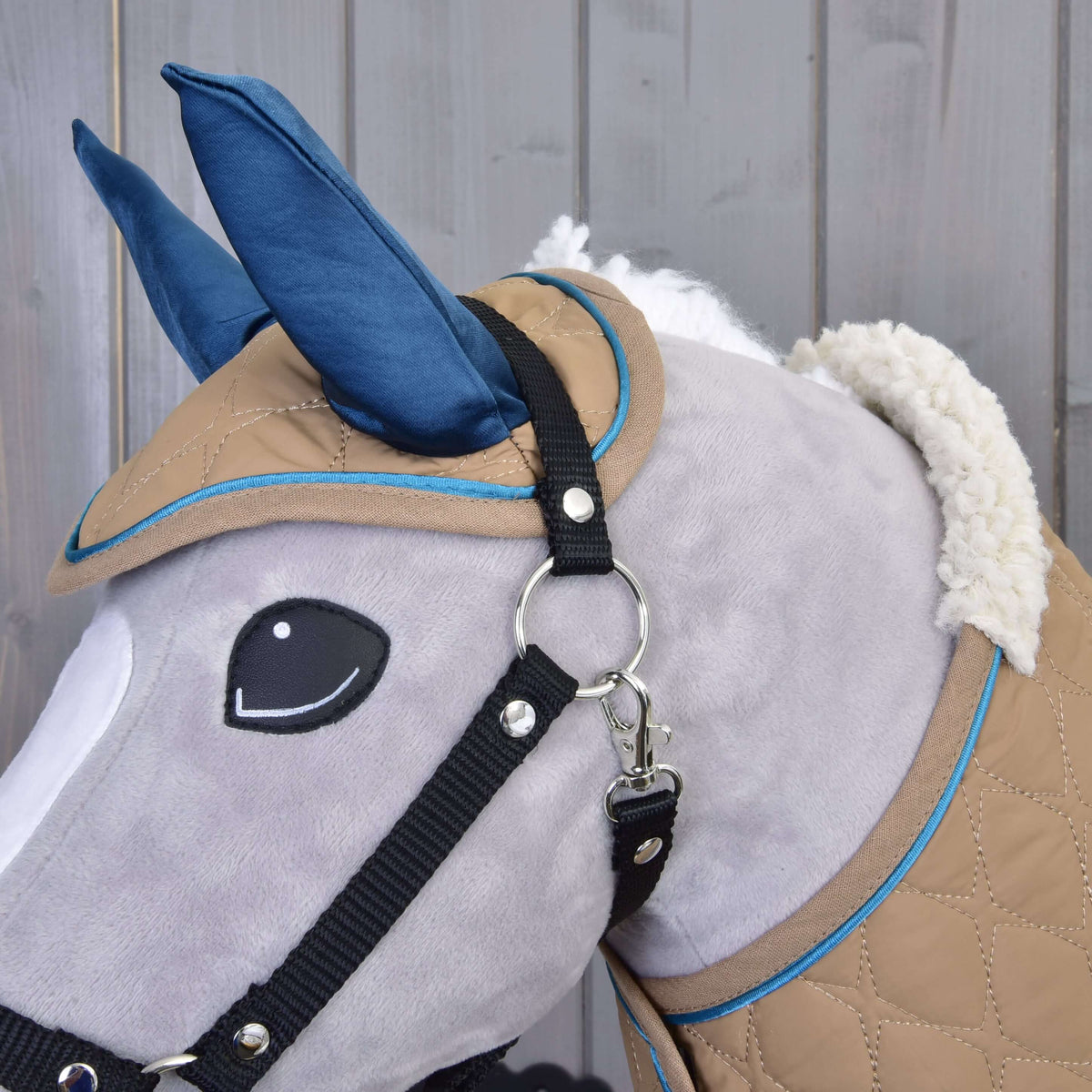 A close-up of the Hobby Horse LarDen Set Almond Star—a gray plush hobby horse with blue ears, black bridle, and tan-blue quilted covering—stands out against a wooden background.