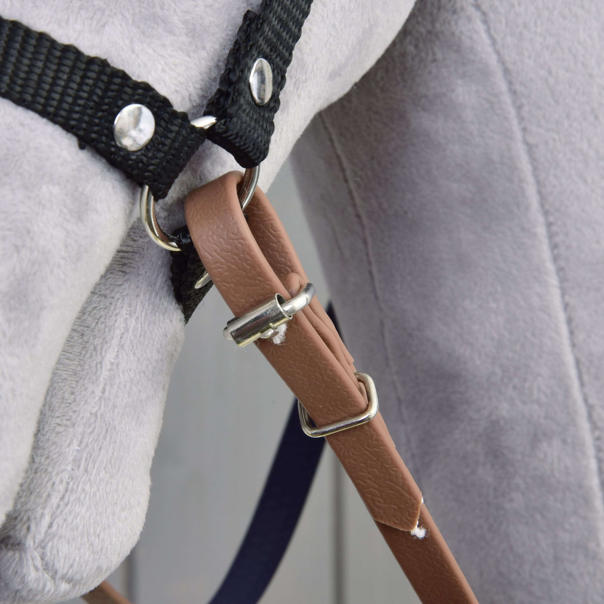 Close-up of a soft, light gray plush toy wearing Hobby Horse LarDen Reins rubberized brown purple—a bitless bridle with black strap harness, metal rings, and brown faux-leather belt.