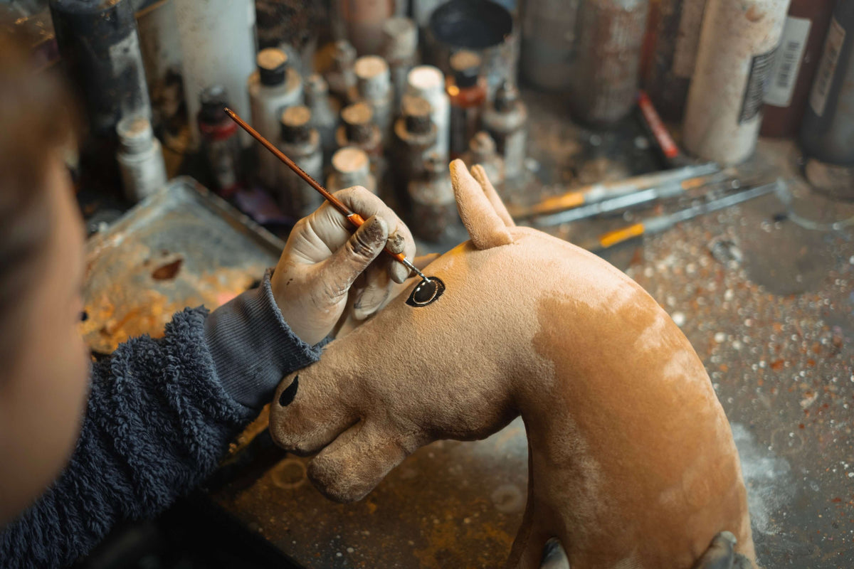 A person wearing gloves paints the eye of a brown Hobby Horse plush toy with a thin brush, surrounded by various paint bottles and art supplies on a cluttered workbench—capturing the creativity of Hobby Horsing.