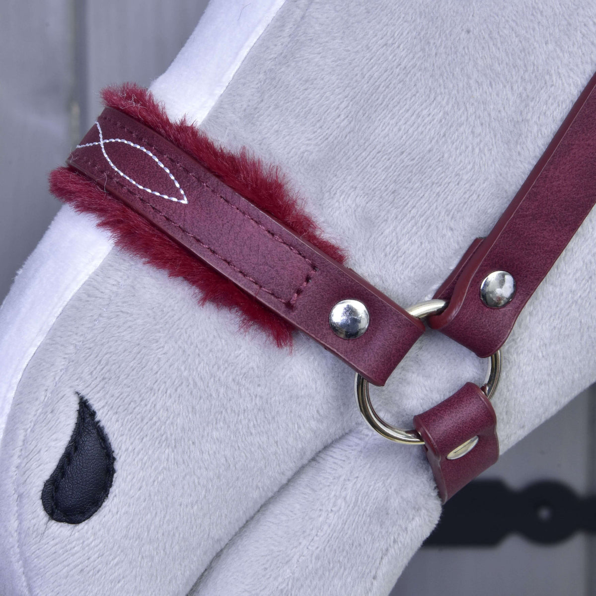 Close-up of a light grey plush horse’s head wearing the Hobby Horse LarDen "Halter with fur - leatherette Burgundy." Features include maroon-padded red leatherette and a black teardrop nostril patch. Ideal as a stylish Hobby Horse accessory. Background is blurred.