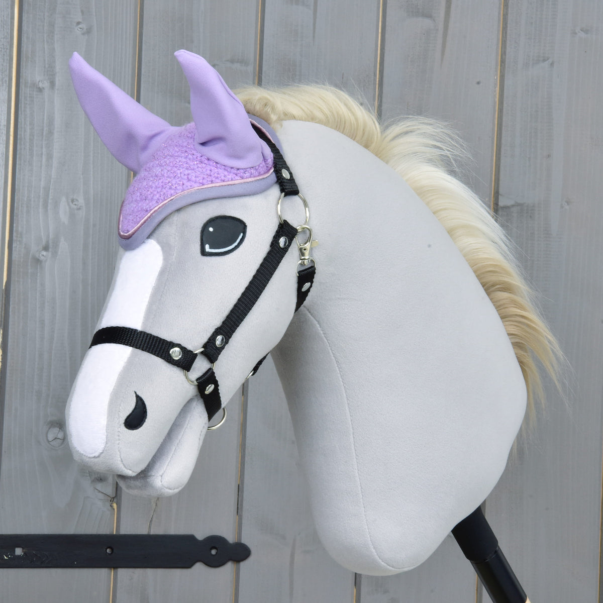 A plush hobby horse with a light gray body, black bridle, and cream mane wears the Hobby Horse LarDen Ear bonnet Orchid for insect protection. The background shows a wooden fence.
