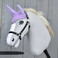 A plush hobby horse with a light gray body, black bridle, and cream mane wears the Hobby Horse LarDen Ear bonnet Orchid for insect protection. The background shows a wooden fence.