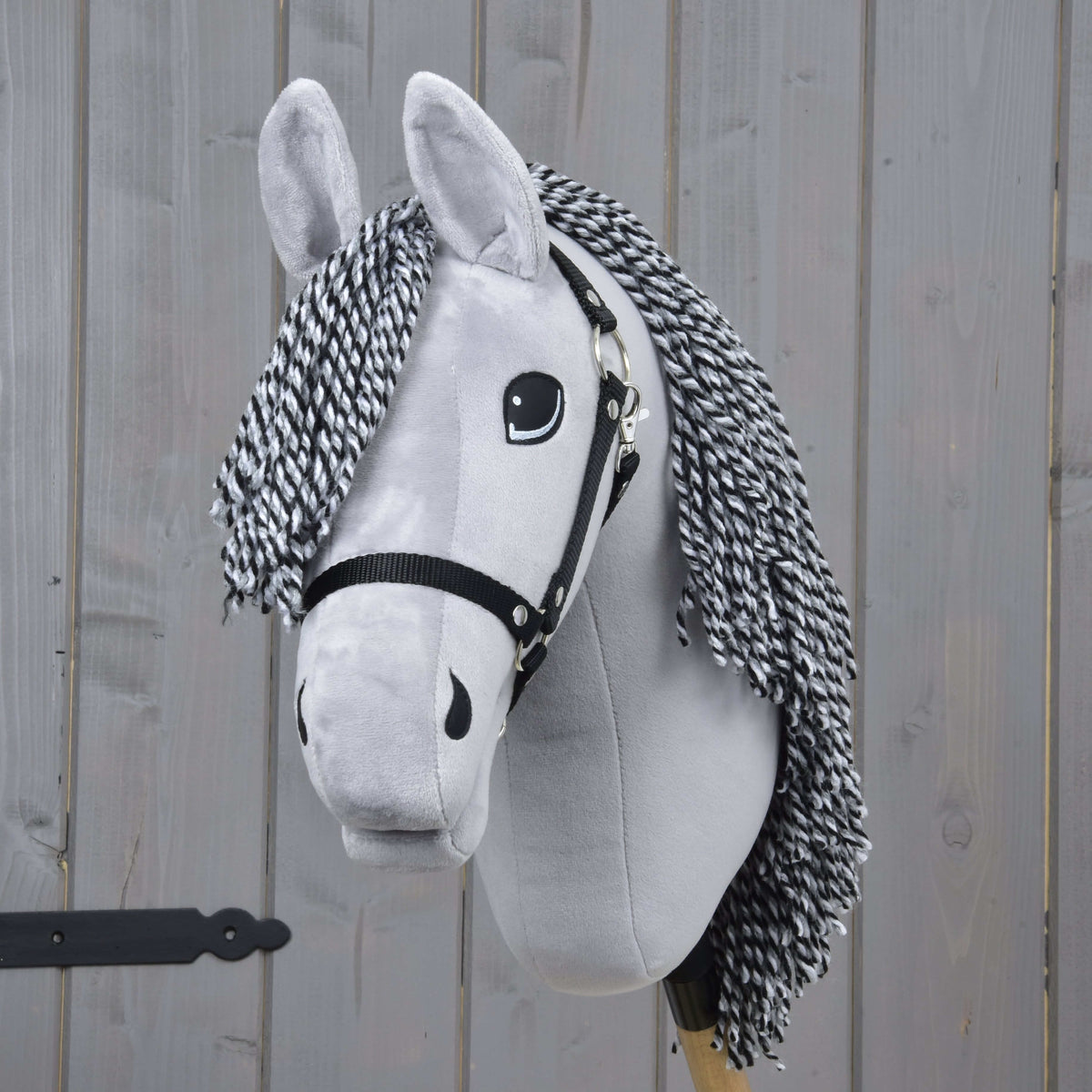 The Hobby Horse Caesar with Black Halter by Hobby Horse LarDen, featuring a gray plush body, black and white yarn mane, black halter, and expressive eyes, is set against a wooden plank background.