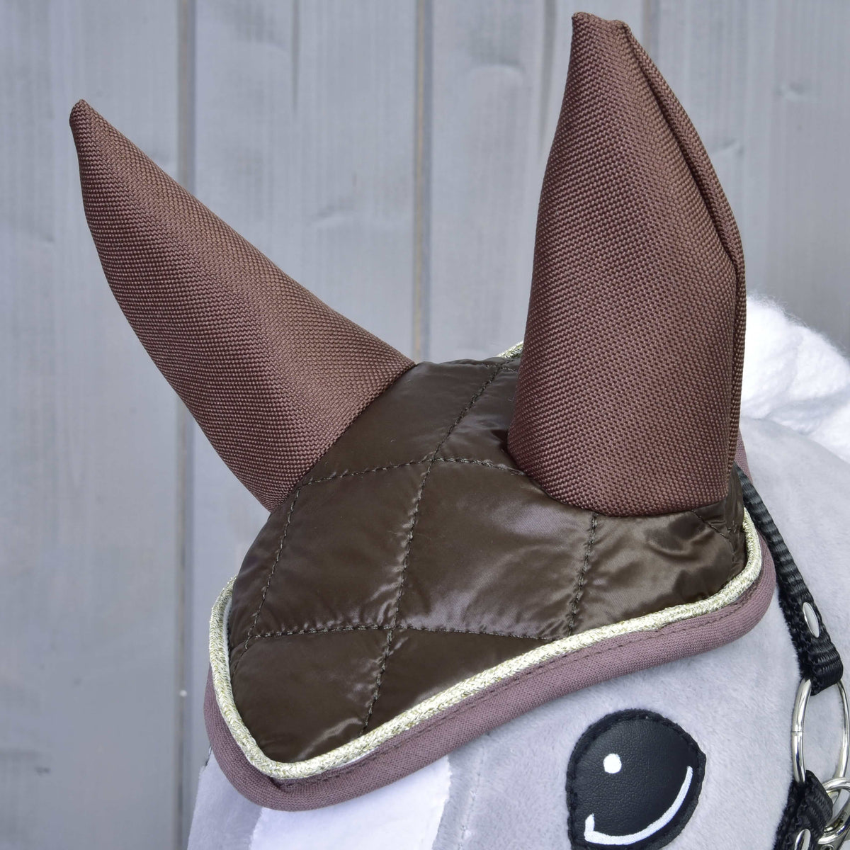 A close-up of the Hobby Horse LarDen Set Thermo Chocolate displays its large fabric ears and quilted brown ear bonnet with light trim—a charming horse decoration against a gray wooden background.