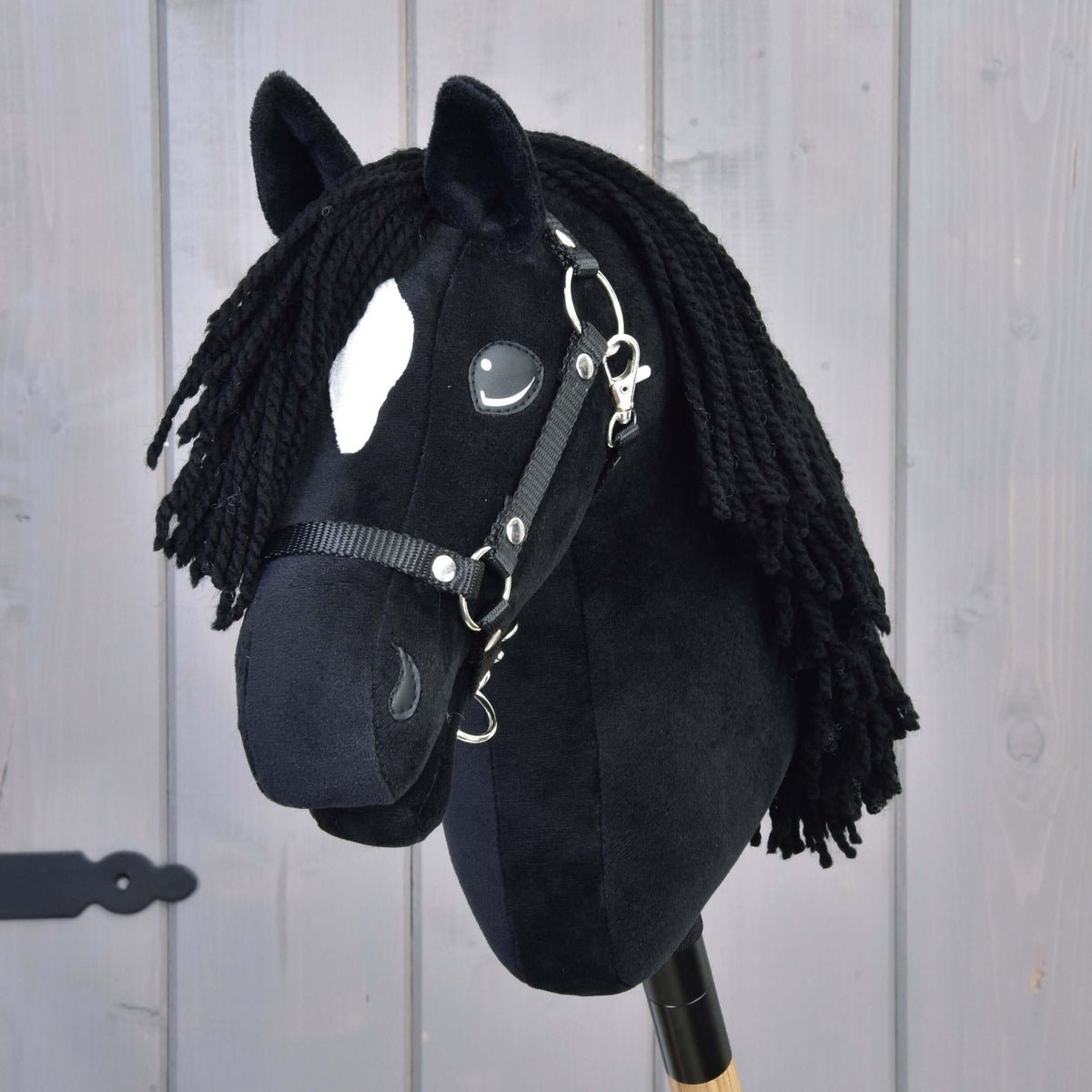 A close-up of the Hobby Horse Cloony with Black Halter by Hobby Horse LarDen, resembling an American Quarter Horse stallion, features a white spot on its forehead, black yarn mane, and a black halter, set against a wooden background.