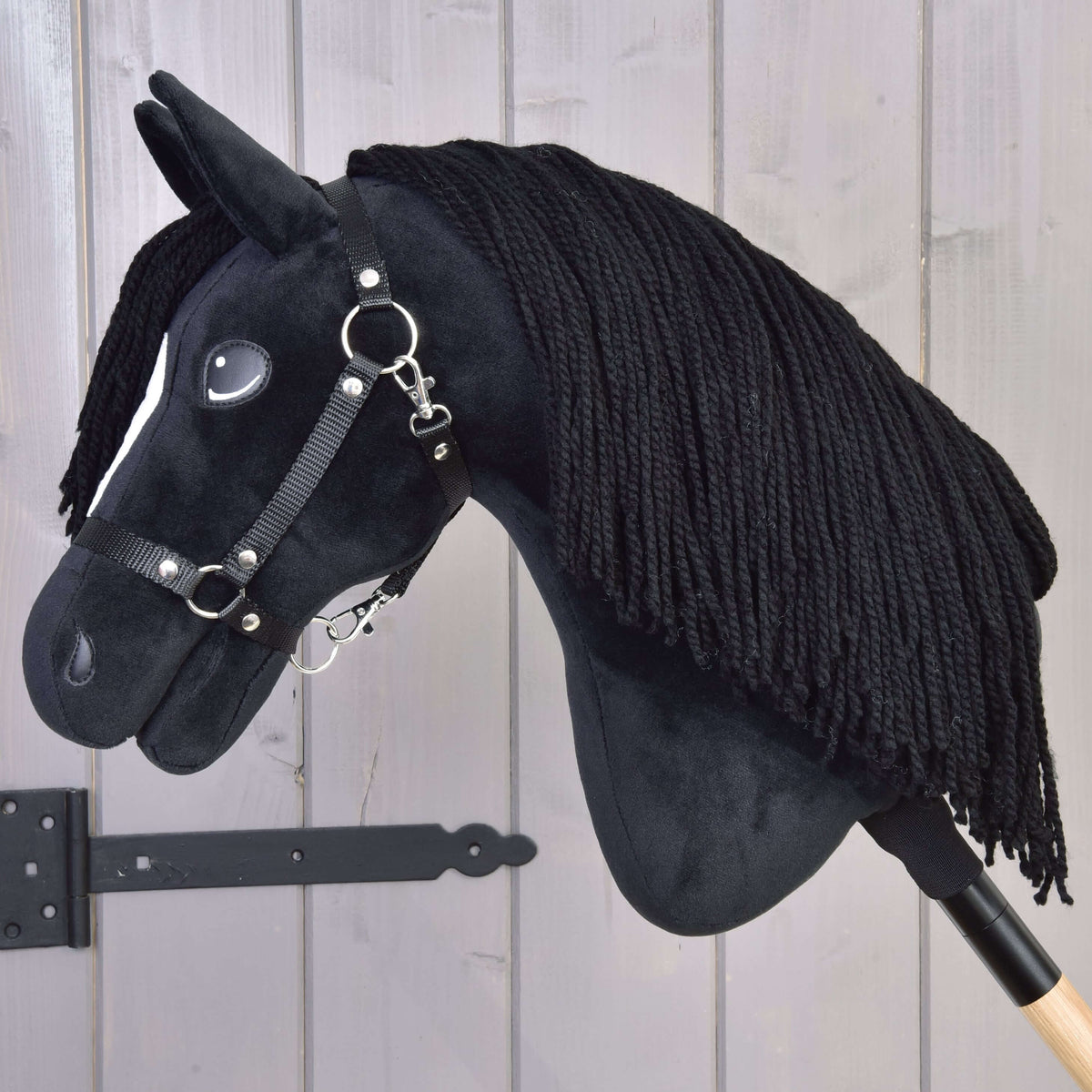 The Hobby Horse Cloony with Black Halter by Hobby Horse LarDen, a black stallion with a white facial spot, black yarn mane, and bridle on a wooden stick, stands before light gray wooden boards.